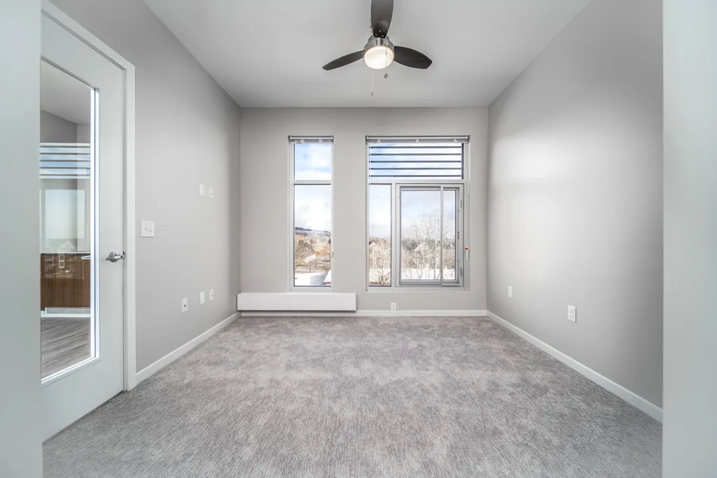 Empty room with large windows, ceiling fan, and neutral gray walls for versatile interior design.