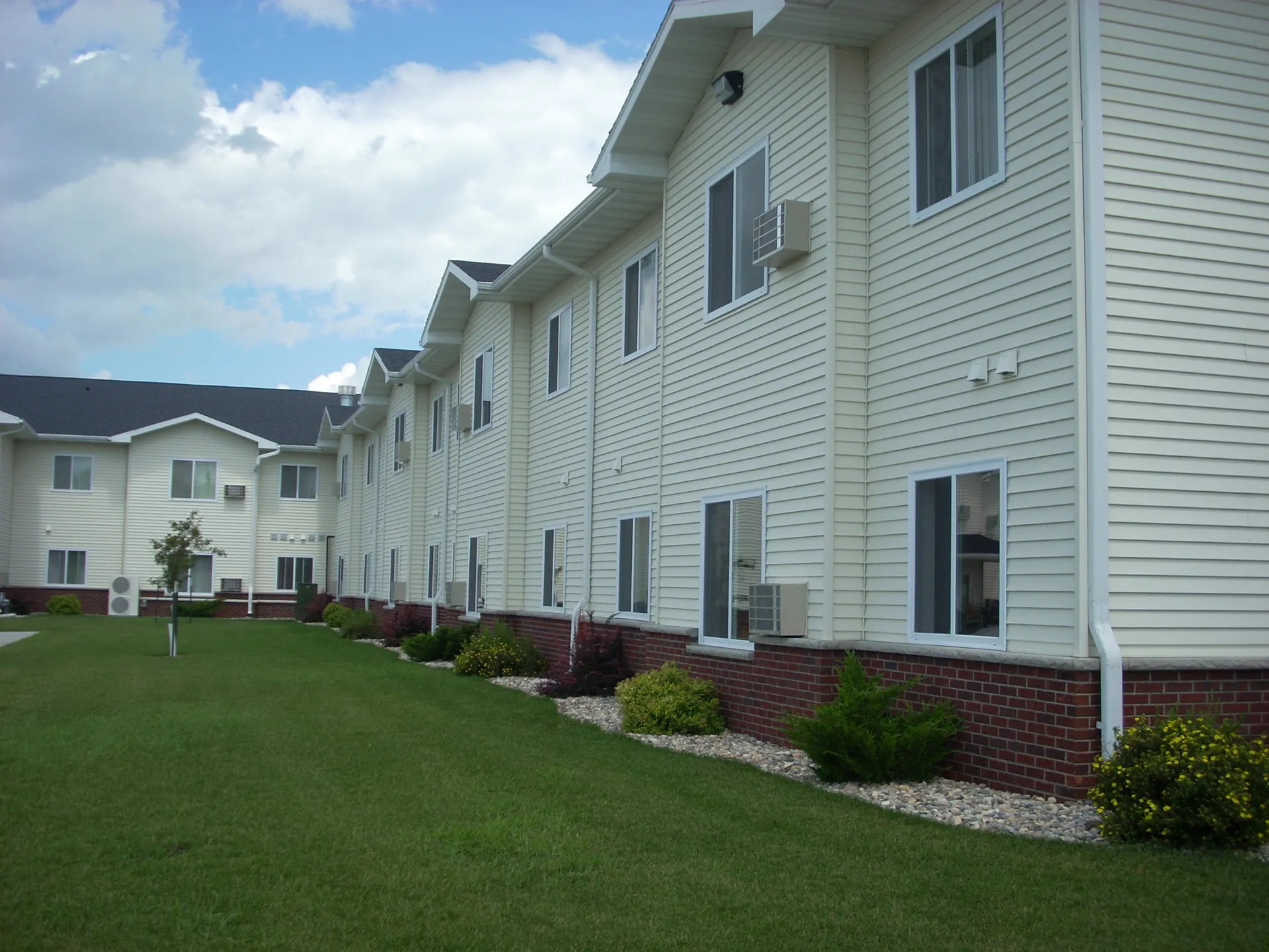 Apartment complex with white siding and brick accents, featuring multiple windows and air conditioni.