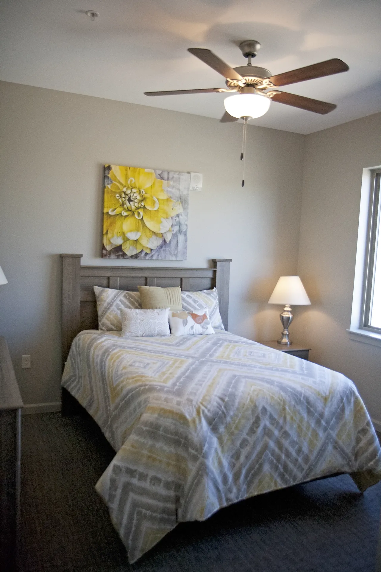 Ceiling fan and natural light illuminate a cozy bedroom with a bed, artwork, and a bedside lamp.