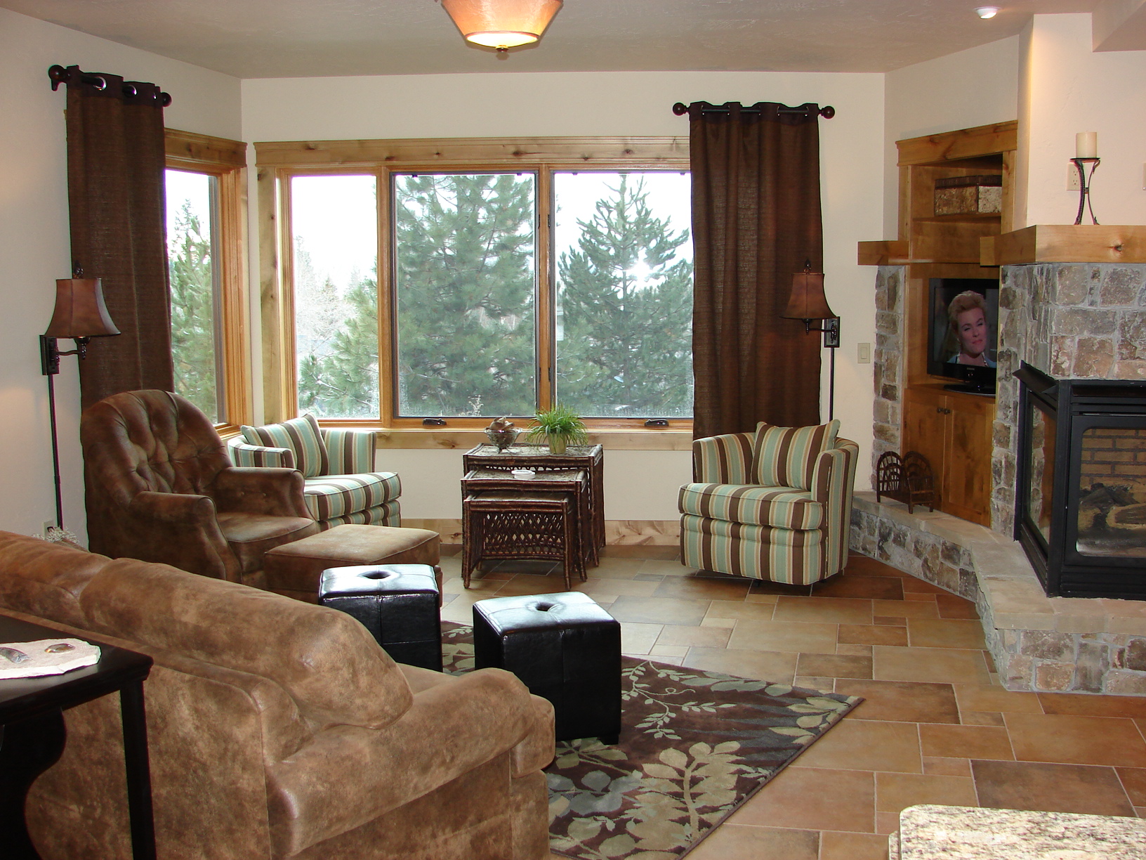 Spacious living room featuring a stone fireplace, large windows with views of trees, and comfortable.