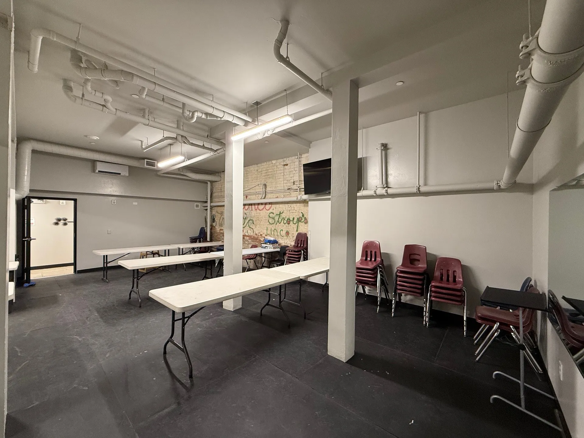 Spacious commercial meeting room with tables and stacked chairs, industrial ceiling, and white walls.