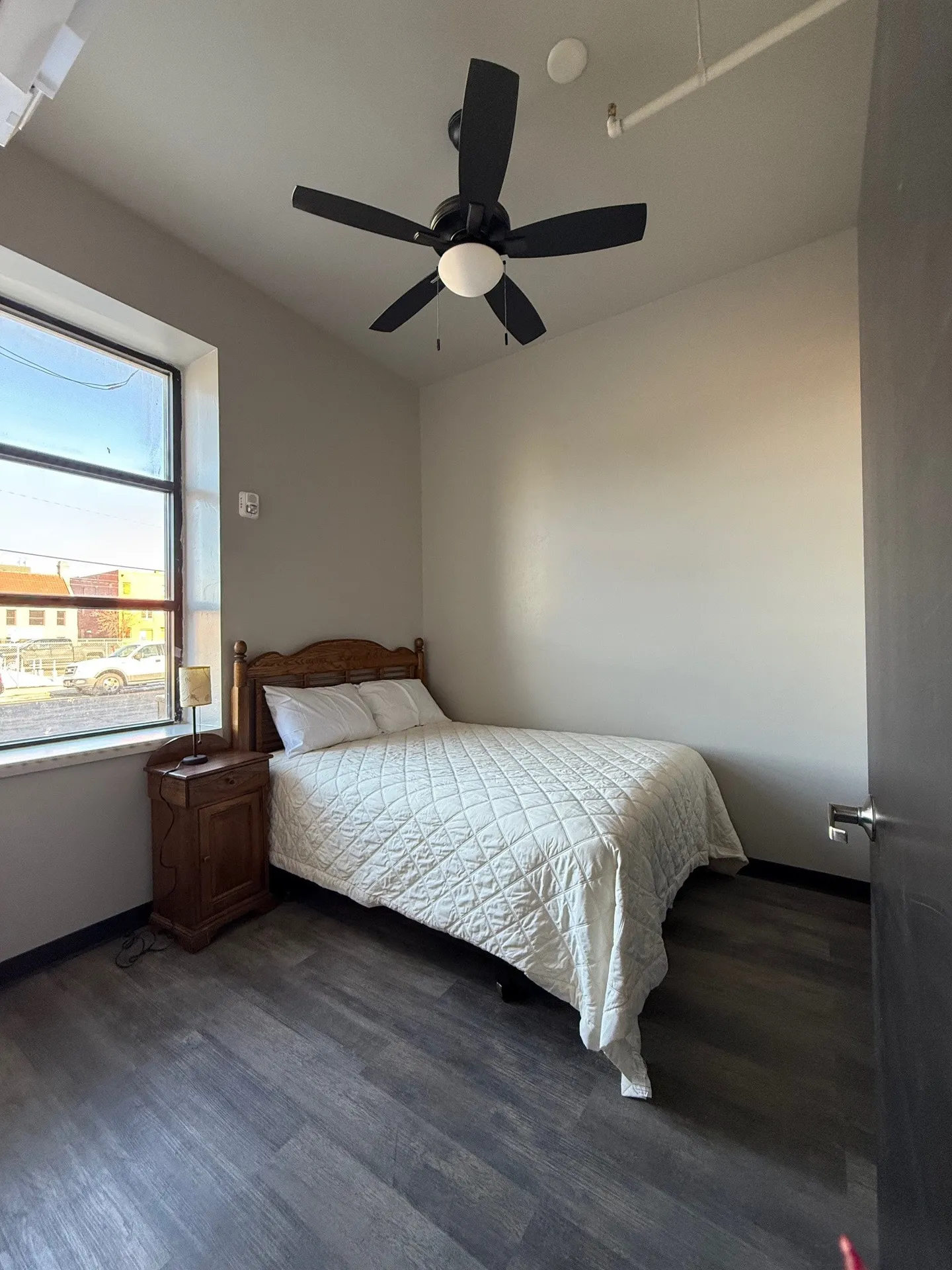 Bright bedroom with large window and ceiling fan, featuring a wooden bed frame and simple decor.