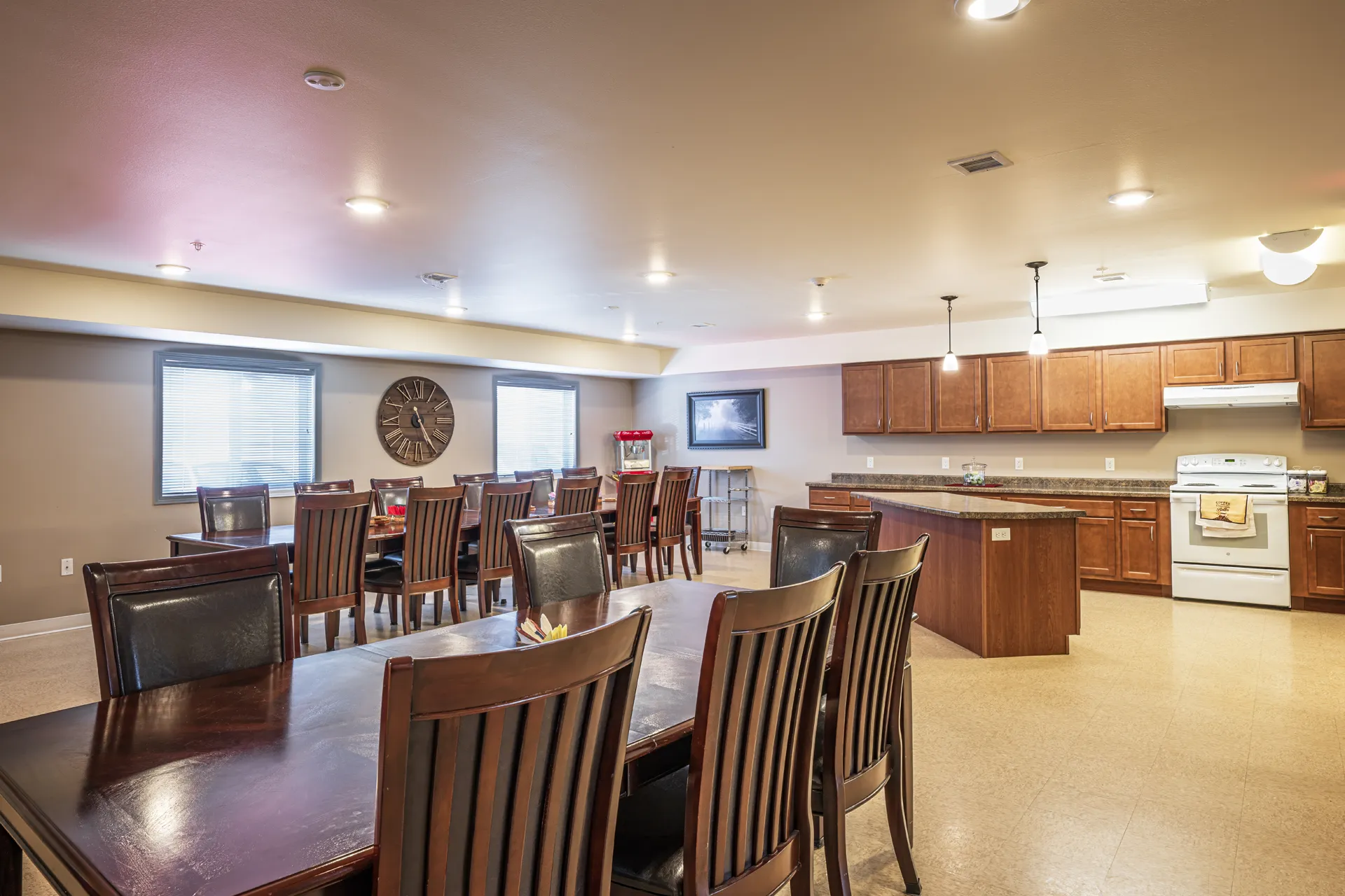 Modern communal dining area with wooden tables and chairs, featuring a kitchen and large windows.